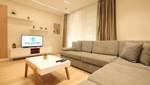 Un salon moderne avec un canapé, une table basse, un téléviseur posé sur un meuble et une étagère murale agrémentée de plantes.