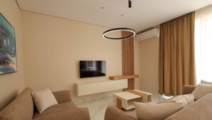 Una sala de estar moderna con una televisión de pantalla plana montada en una consola blanca, una lámpara de techo redonda y un amplio ventanal con cortinas.