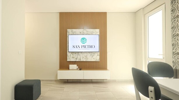 Une pièce moderne et minimaliste, dotée d’un téléviseur à écran plat fixé sur un mur recouvert de panneaux en bois, d’une petite table blanche et de deux chaises noires.