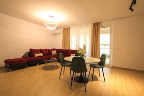 Un salon moderne avec un canapé d’angle, une table de salle à manger ronde et des chaises. On y trouve un luminaire au plafond et une fenêtre avec des rideaux.