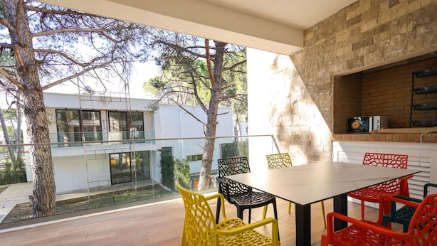 A modern outdoor dining area with a glass balustrade, a brick wall, and a view of a contemporary building. - Sanpietro Vacation Apartment 030 (Ishëm)
