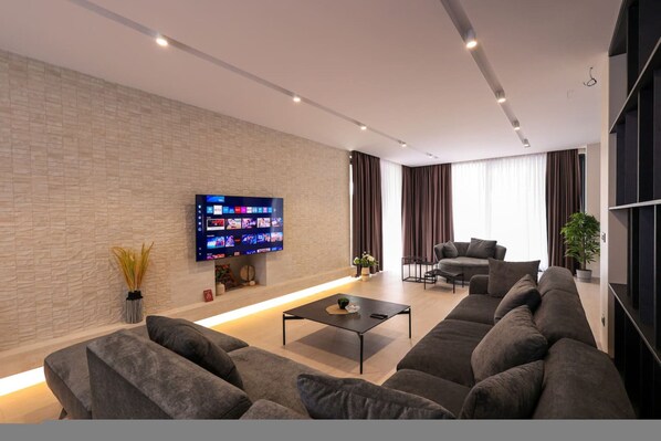 A modern living room with a large flat-screen TV mounted on the wall, a sectional sofa, a coffee table, and a potted plant.