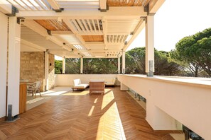 A covered outdoor area with a herringbone floor, white walls, and a stone wall on one side. There are two white sofas and a wicker chair. - Sanpietro Elite Signature Villa (Ishëm)