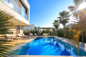 A modern pool area with lounge chairs and a covered seating area, surrounded by palm trees and a clear sky. - Sanpietro Vacation Villa 005 (Ishëm)