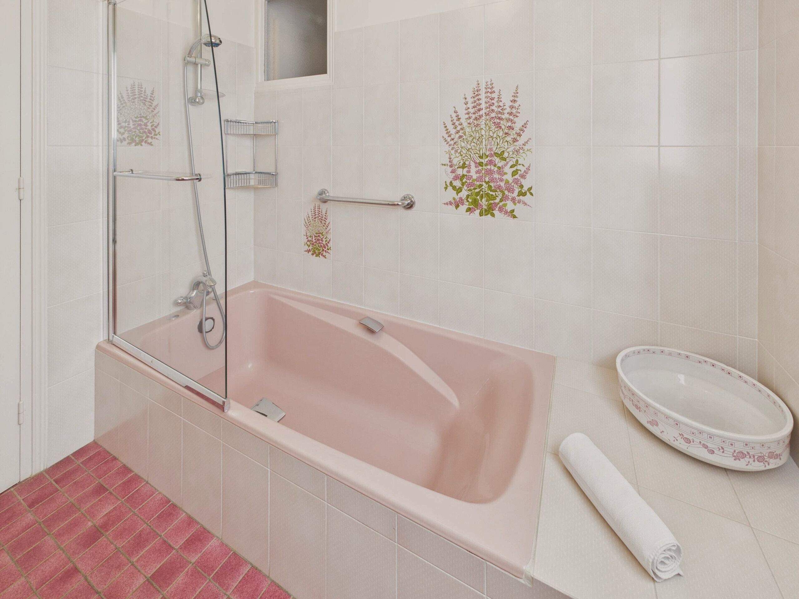 Uma casa de banho com uma banheira rosa, um chuveiro com divisória de vidro, uma pia branca com design floral e uma toalha branca no chão.