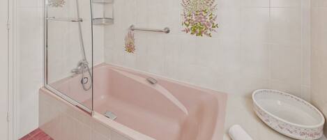 Uma casa de banho com uma banheira rosa, um chuveiro com divisória de vidro, uma pia branca com design floral e uma toalha branca no chão.
