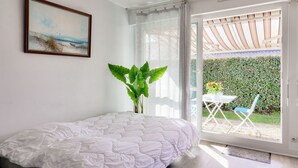 A bedroom with a bed, a window with a view of a garden, a painting on the wall, and a potted plant.