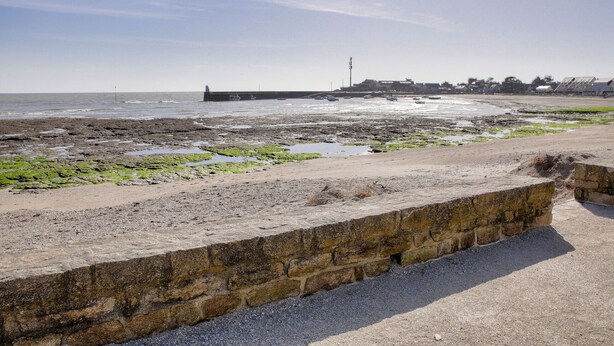 A coastal area with a stone wall, a beach, and a pier in the background.