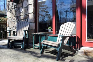 Deux fauteuils Adirondack sont placés sur une terrasse en bois, à l’extérieur d’un bâtiment doté d’un mur en pierre et d’une porte rouge.