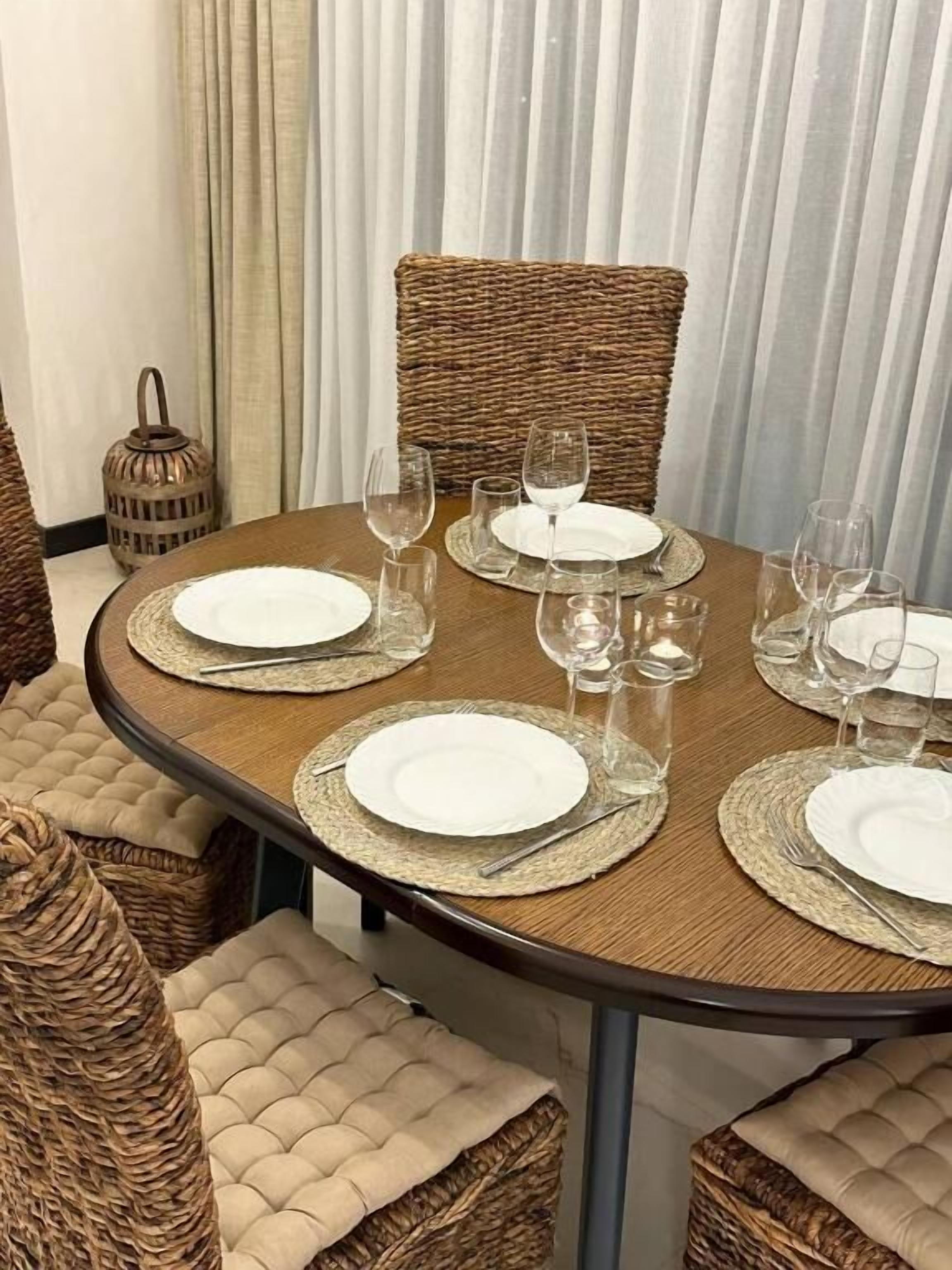 Una mesa de comedor redonda con platos blancos, vasos transparentes y individuales tejidos.