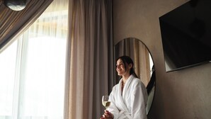 Iemand in een witte badjas zit op een bankje in een moderne hotelkamer met een grote spiegel, een televisie en uitzicht op het buitengebied.