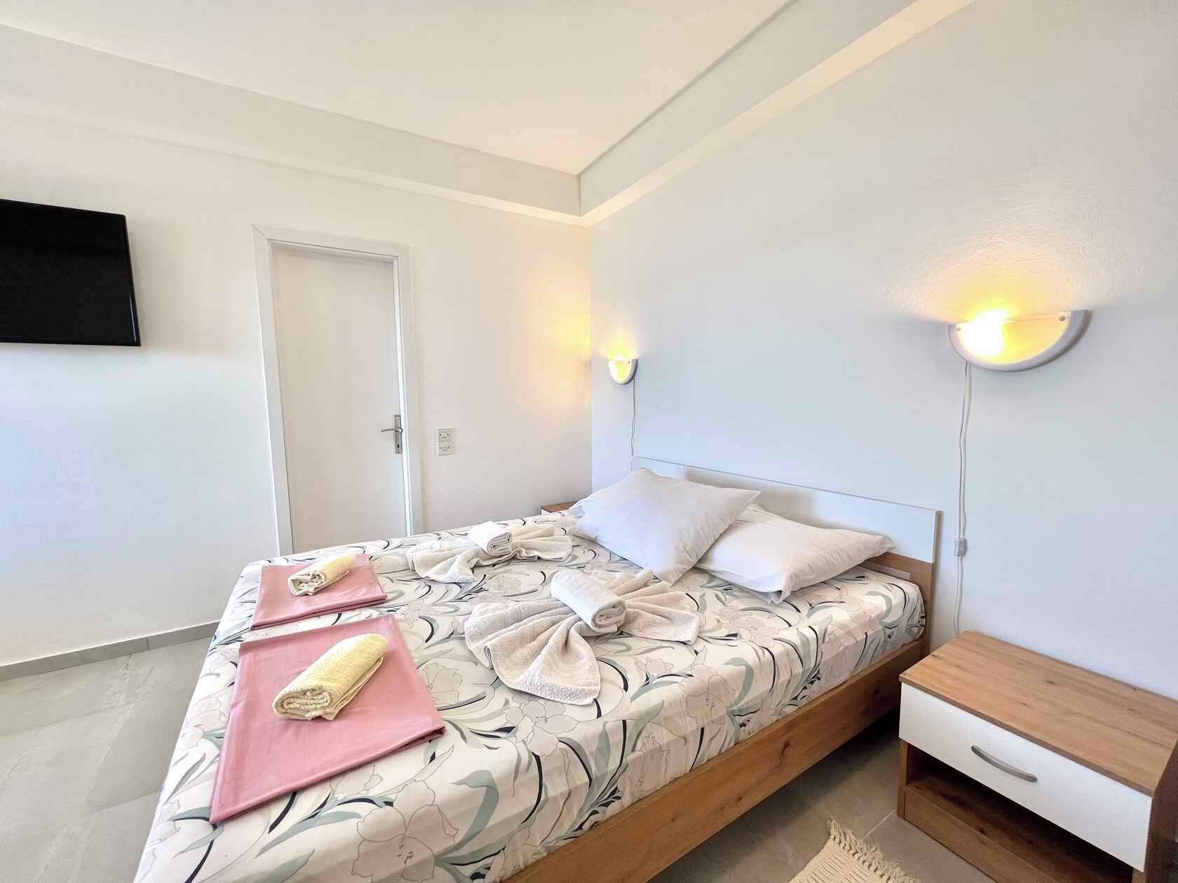 Una cama bien hecha con toallas, una mesita de noche con lámpara y un televisor en la pared.