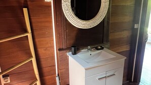 Un bagno con pavimento in legno, un lavandino bianco, uno specchio con cornice strutturata e una scala di legno appoggiata al muro.