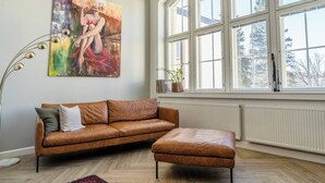 Un salon moderne avec un canapé en cuir marron, un pouf assorti, une lampe sur pied et une grande fenêtre donnant sur des arbres.