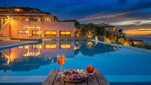 A swimming pool with a wooden table in the foreground, a building with warm lighting in the background, and a sunset sky. - Le Dalie With Shared Pool (Olbia)