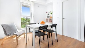 Una habitación luminosa y de color blanco, con piso de madera, una mesa blanca, sillas negras, una silla gris y una ventana con vista a vegetación.