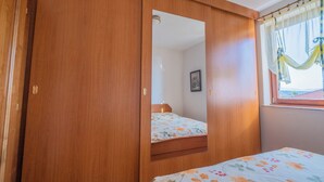 Ein Schlafzimmer mit Holzschrank, einem Bett mit floralem Bettzeug, einem Fenster mit transparenten Vorhängen und Blick ins Freie.