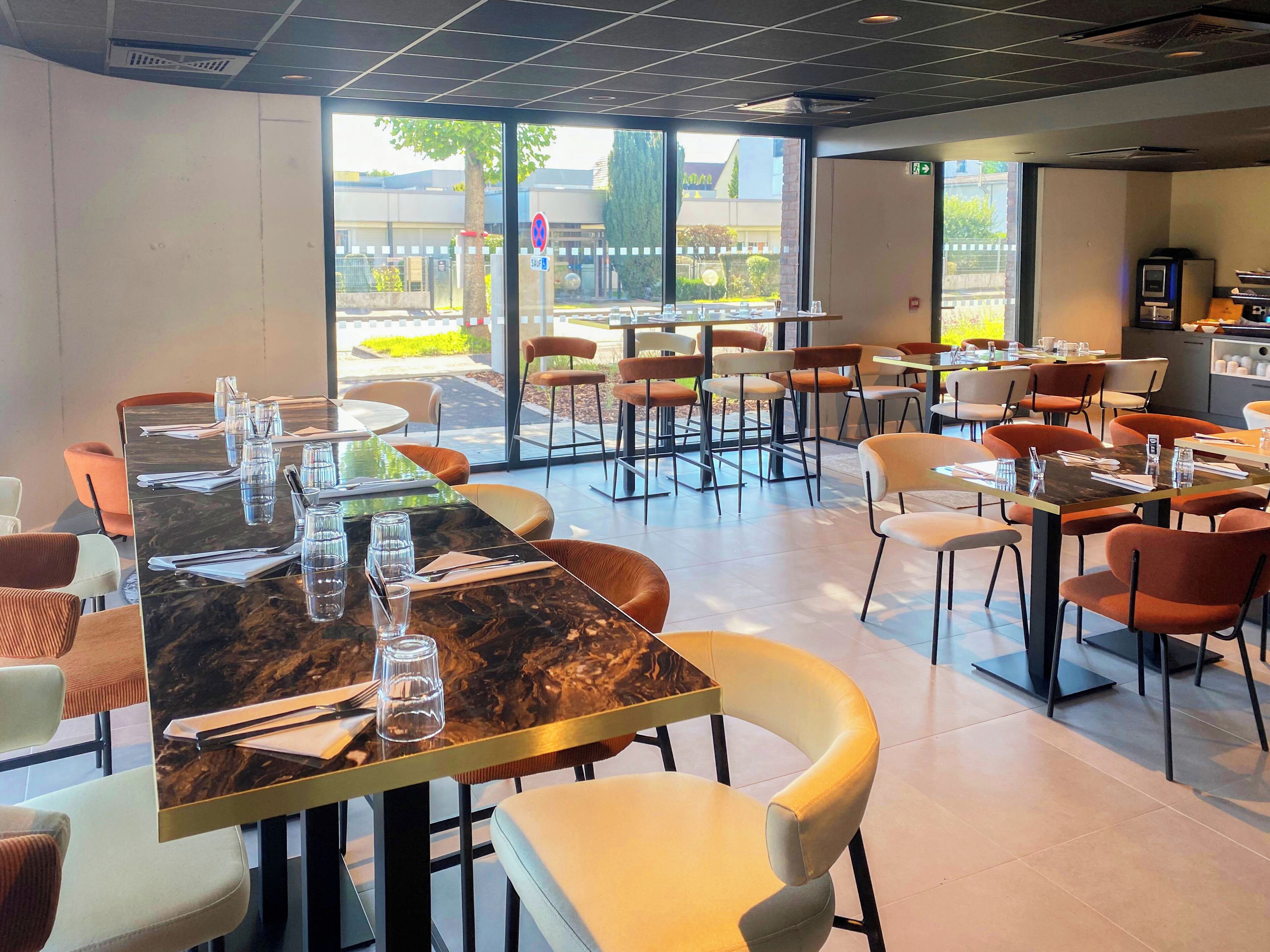 A modern dining area with marble tables and cushioned chairs, large windows offering a view of the outdoors, and a clean, contemporary design.