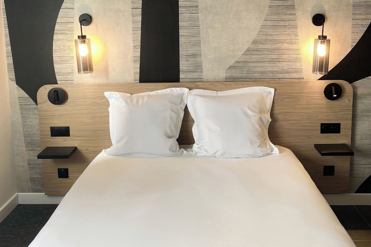 A modern hotel room with a wooden headboard, two white pillows, and wall-mounted lighting.