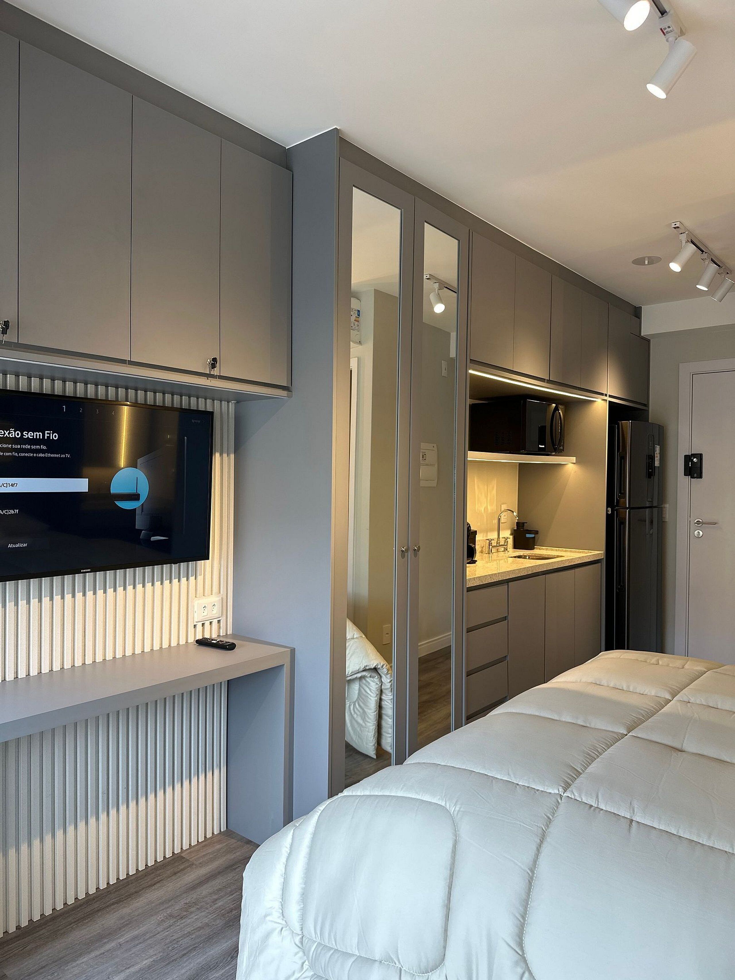 Ein modernes Schlafzimmer mit einem großen Bett, einem an der Wand montierten Fernseher und einem begehbaren Kleiderschrank mit Spiegel.