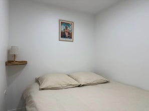 Een slaapkamer met een bed, twee kussens, een houten plank met een lamp en een poster aan de muur.