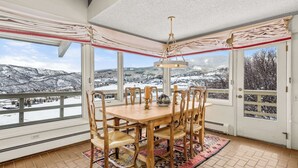 Une salle à manger avec une grande table et des chaises, un lustre, et une vue sur un paysage montagneux enneigé, visible à travers de grandes fenêtres.
