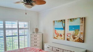 Un dormitorio con cama, cómoda, ventilador de techo y dos cuadros con motivos playeros en la pared.