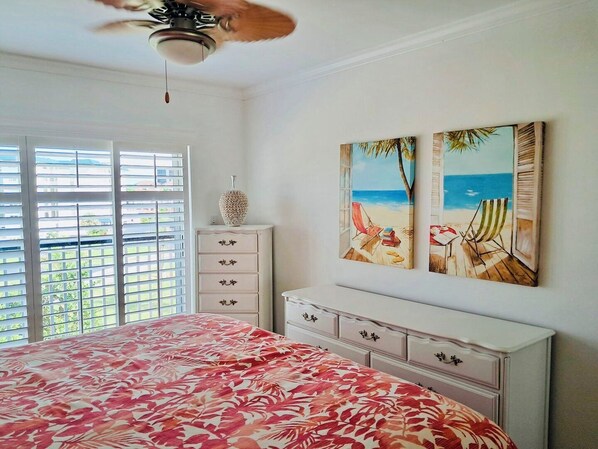 Un dormitorio con cama, cómoda, ventilador de techo y dos cuadros con motivos playeros en la pared.