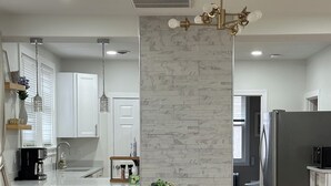 Una cocina moderna con armarios blancos, salpicadero de mármol y un comedor con mesa de madera y sillas de mimbre.