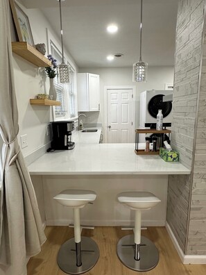 Una cocina moderna con armarios blancos, una isla central con taburetes, lavadora integrada e iluminación colgante.