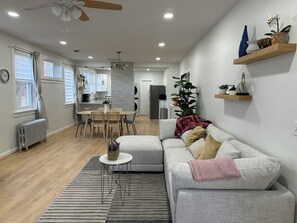 Una sala moderna con un sofá gris, una mesa de centro y un tapete a rayas. Se aprecia una zona de comedor con una mesa y sillas, y una cocina al fondo.