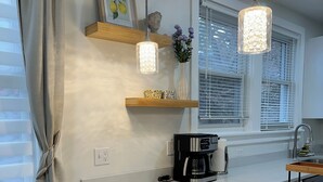Una cocina moderna con encimera blanca, dos taburetes de bar, cafetera y lámparas colgantes.