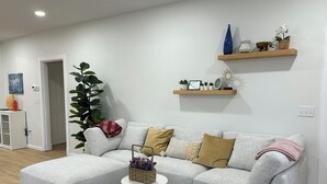 Una sala moderna con un sofá seccional gris, una mesita de centro redonda y una planta grande en maceta. Dos estantes de madera sobre el sofá exhiben objetos decorativos.