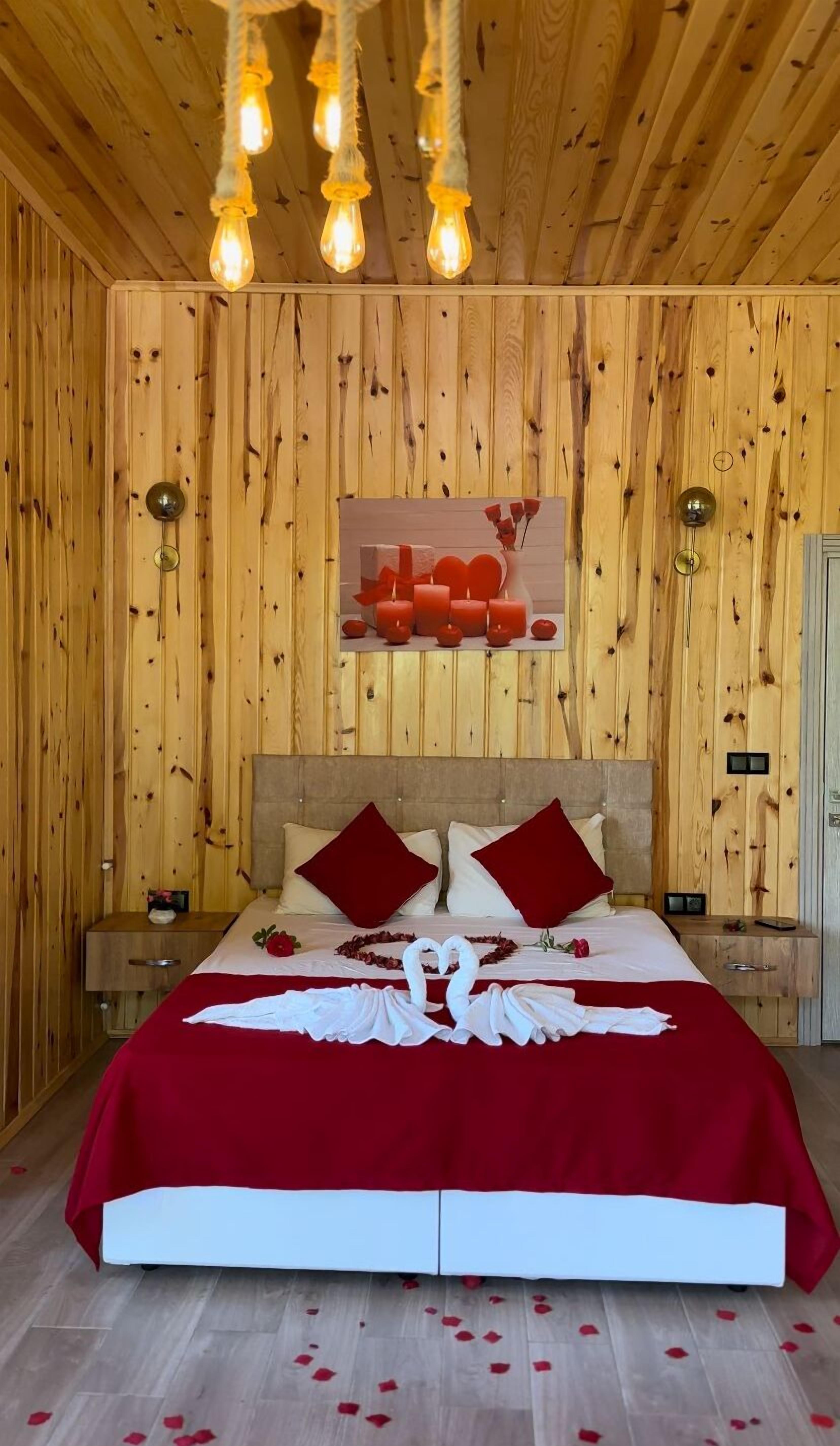 Un dormitorio con paneles de madera, una cama, almohadas rojas, una disposición de toallas en forma de corazón y una lámpara fijada a la pared.