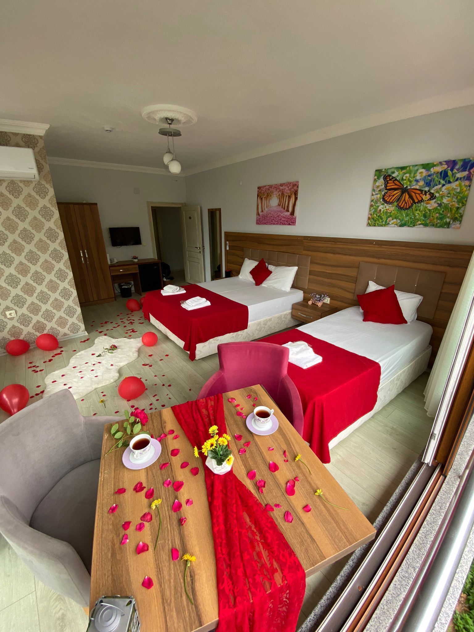 Habitación de hotel con dos camas, una mesa de comedor con pétalos de rosa y un mantel individual rojo, y un televisor en la pared.