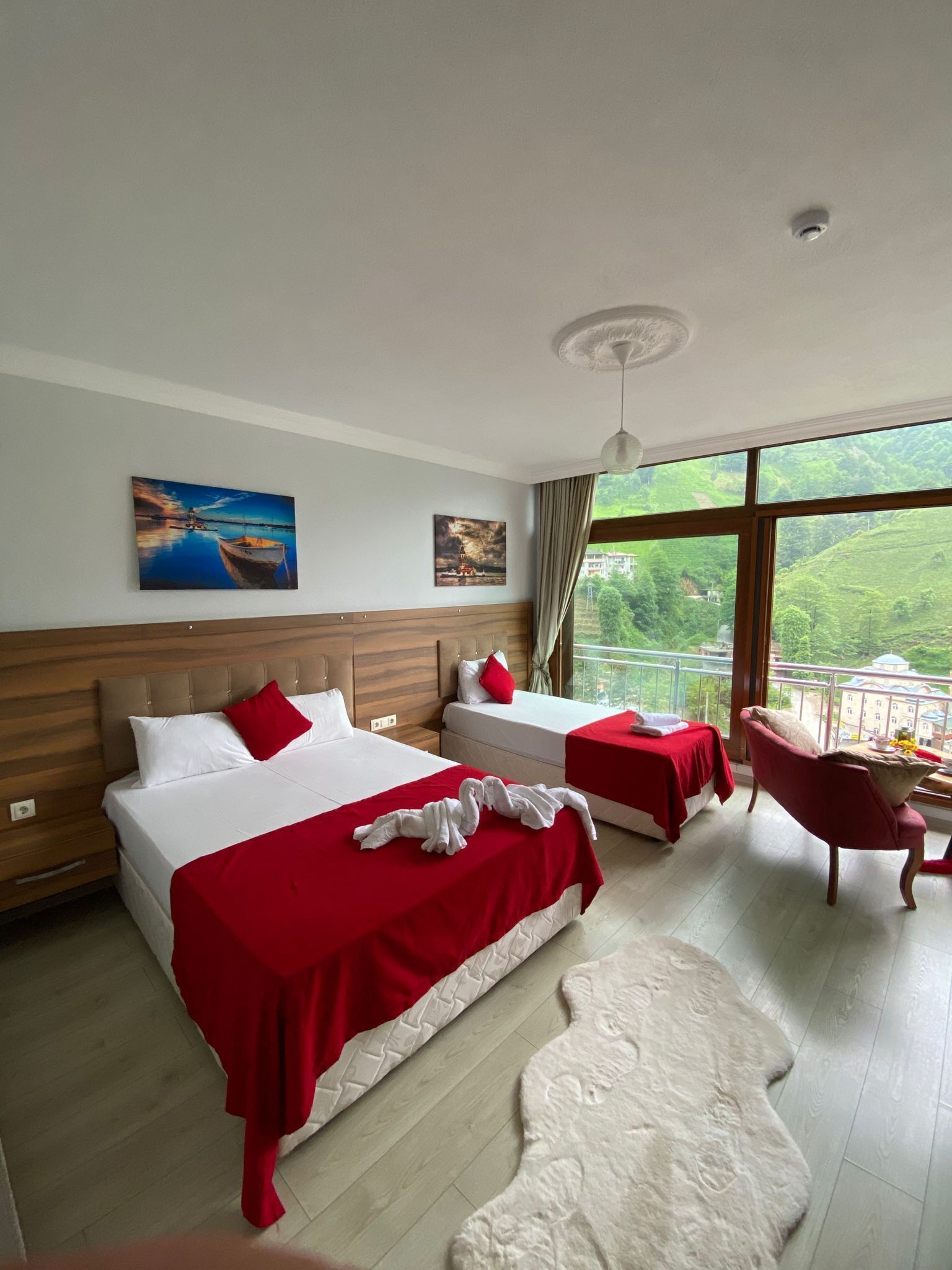 A hotel room with two beds, a red and white color scheme, a large window with a view of greenery, and a small balcony.