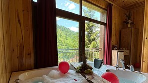 Una cabaña de madera con una bañera grande llena de burbujas y dos bolas rojas flotando, una mesa de madera con frutas, y vistas a colinas verdes a través de grandes ventanales.