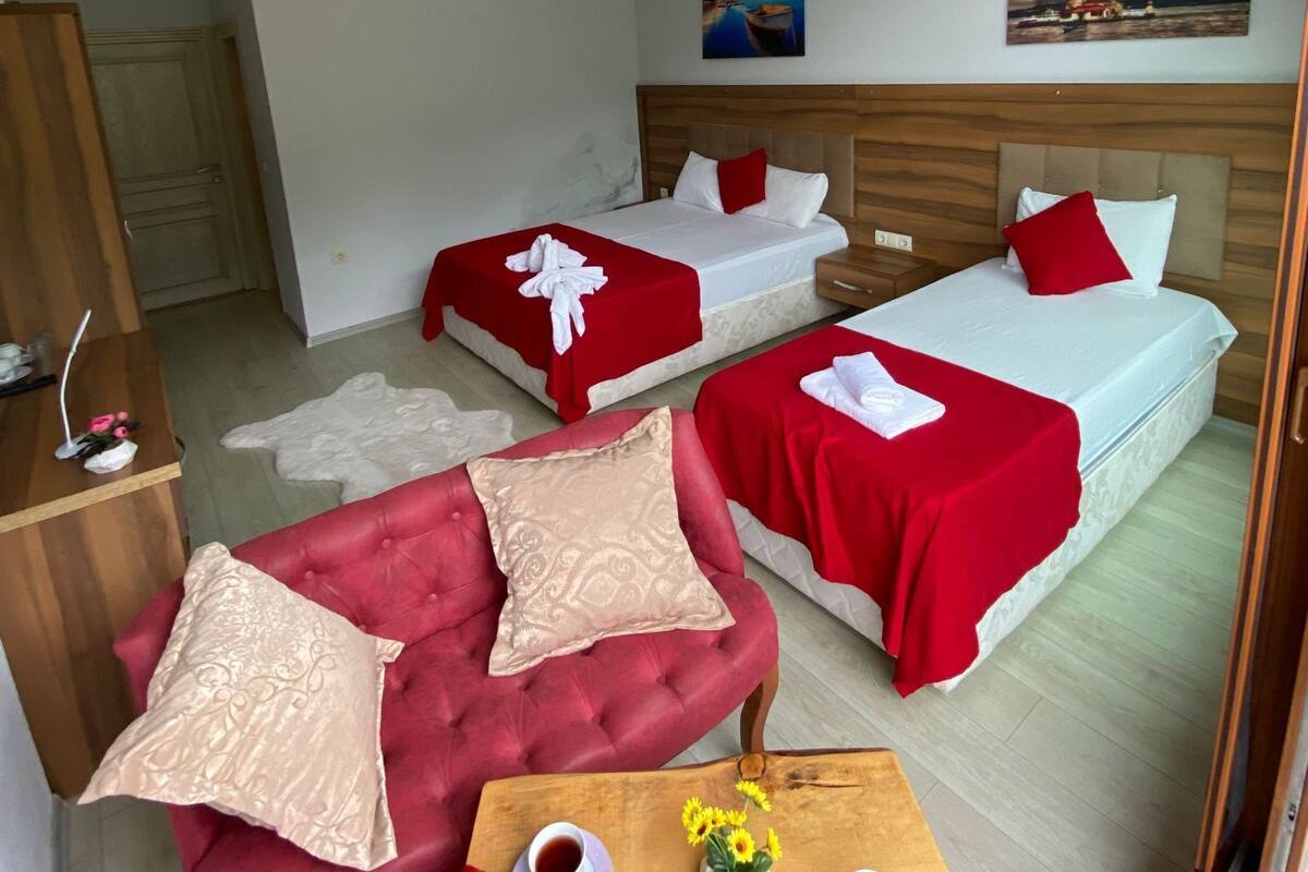 Une chambre d’hôtel avec deux lits, un canapé rouge, une table en bois avec des tasses et des fleurs, et un tableau accroché au mur.