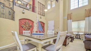 A spacious living area with a dining table, white chairs, a sofa, and a staircase with ornate railings. - Pool, Movie Room, Games, Grill! (Goodyear)