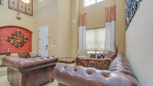 A spacious living room with a brown leather sofa, a red accent wall, and a large window with curtains. - Pool, Movie Room, Games, Grill! (Goodyear)