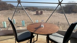 A dining area with a round table and two chairs, a glass vase, and a view of a natural landscape through a geometric window structure.