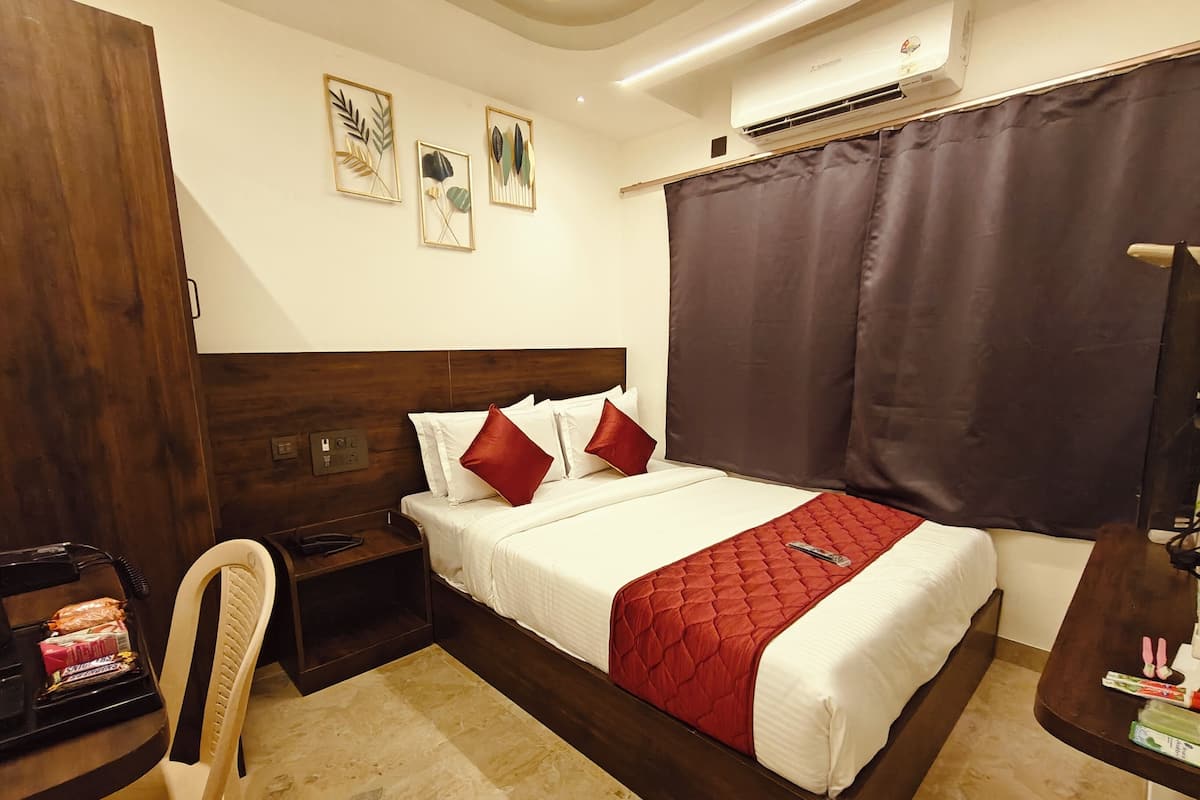 A hotel room with a wooden headboard, a bed with white bedding and red pillows, a bedside table with a lamp, a chair, and a small desk with snacks.