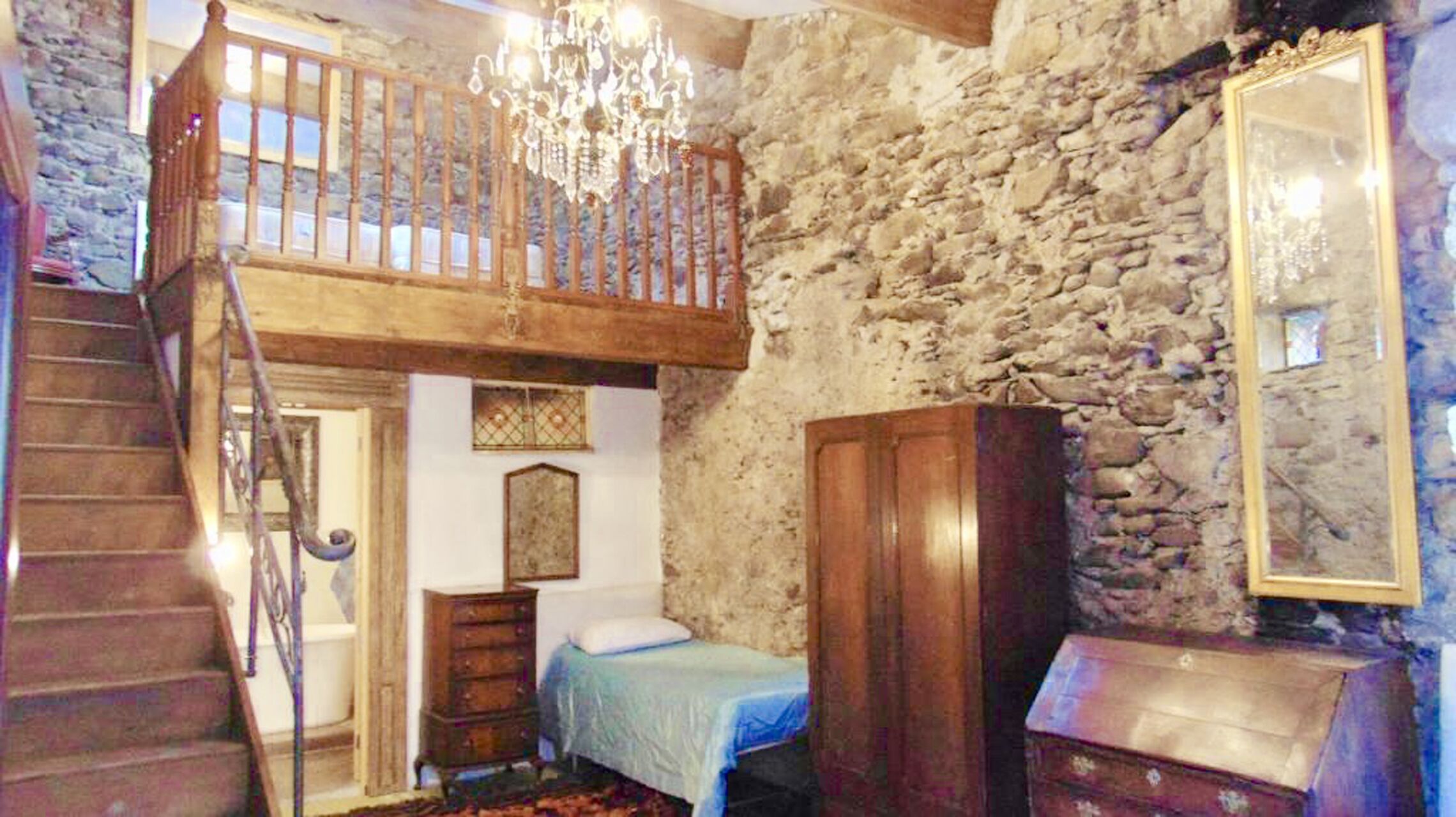 A rustic room with a wooden staircase, a chandelier, a bed, a dresser, and a stone wall.
