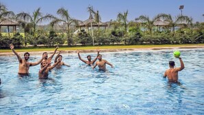 A swimming pool with people playing with a ball, surrounded by palm trees and resort-style buildings. - MST Farm & Resort (Ghaziabad)