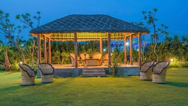 A gazebo with a thatched roof, surrounded by chairs and a small table, set in a well-maintained garden with grass and plants. - MST Farm & Resort (Ghaziabad)