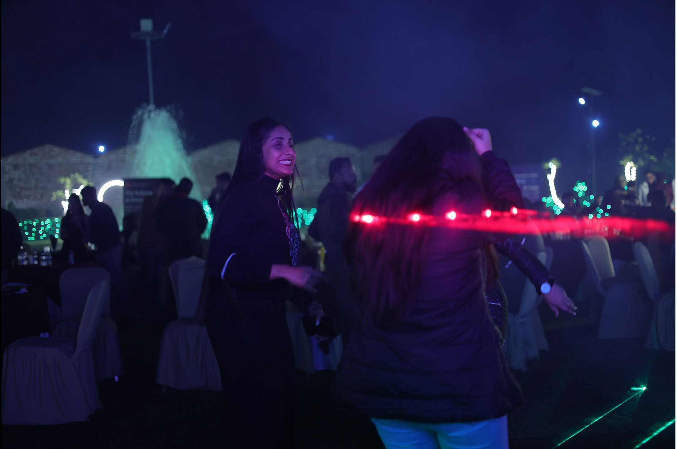 Un grupo de personas bailando en un evento al aire libre, con una fuente y luces decorativas de fondo.