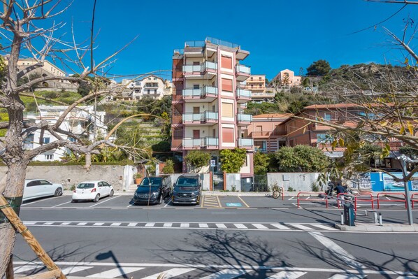 A multi-story residential building with balconies, a parking area with cars, and a clear blue sky.