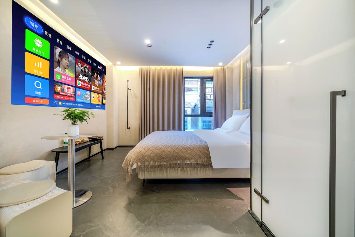 Ein modernes Hotelzimmer mit einer großen digitalen Wand, einem Bett mit beiger Bettdecke, einem kleinen runden Tisch mit Pflanze und Blick auf die Stadt durch das Fenster.