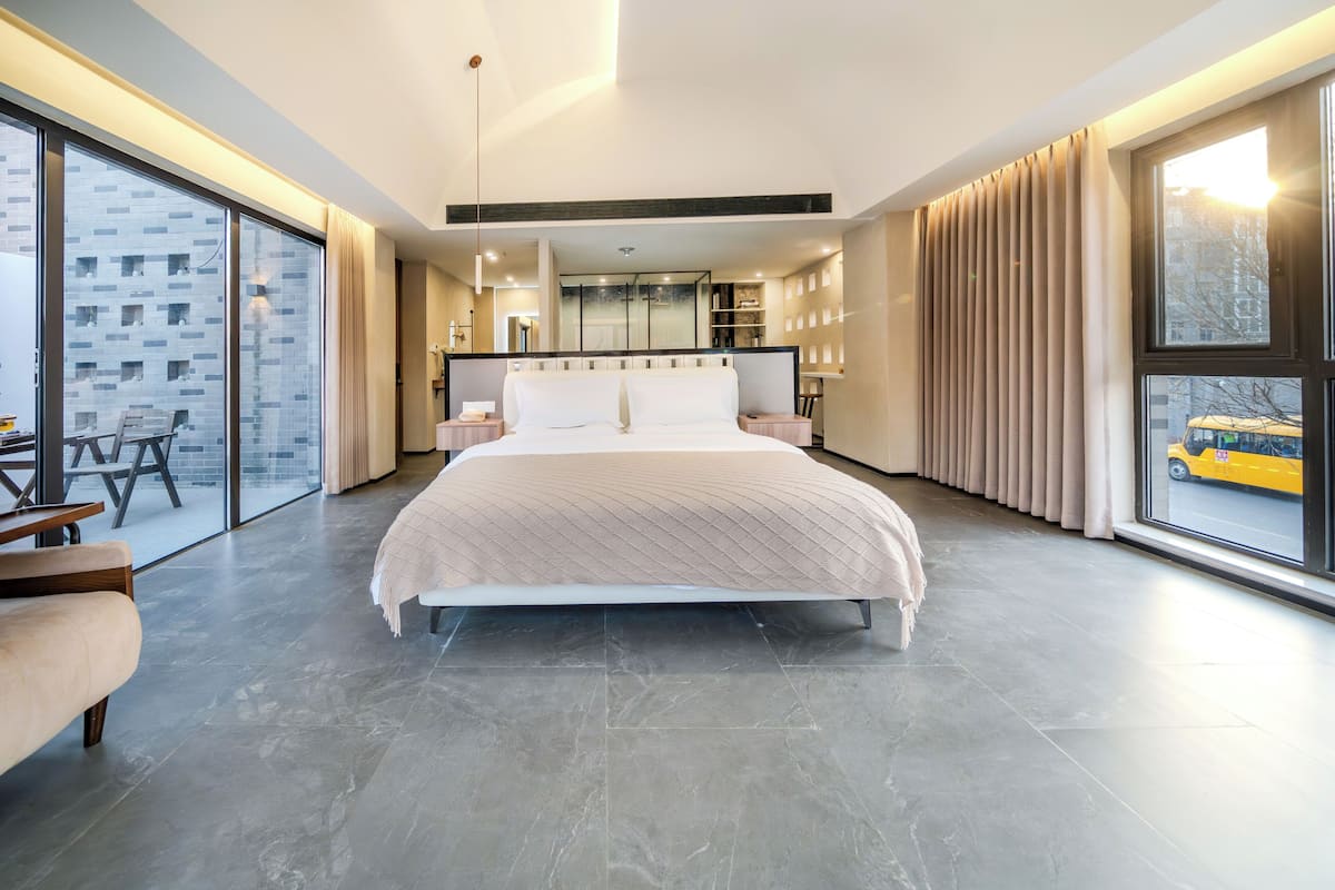 Ein modernes Schlafzimmer mit einem großen Bett, einem Schreibtisch und Blick auf eine Stadtlandschaft durch große Fenster.