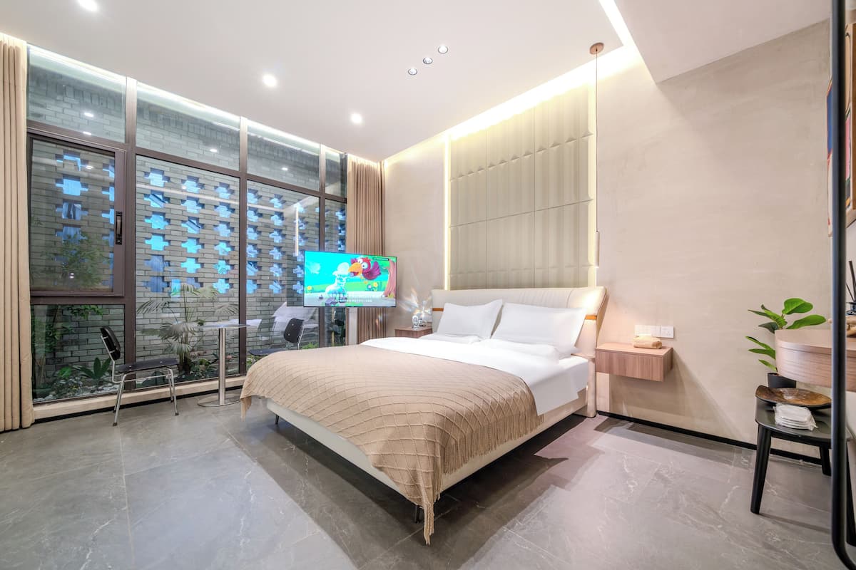 Ein modernes Hotelzimmer mit einem großen Bett, einem Fernseher, einem Nachttisch und einem Blick auf die Stadt durch große Fenster.